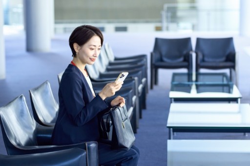 一位穿著深色西裝的商務女性坐在現代化候機室中查看手機，手中同時提著手提包，背景為整齊排列的座椅和玻璃桌子，環境明亮寬敞，展現現代商務旅行的專業形象。